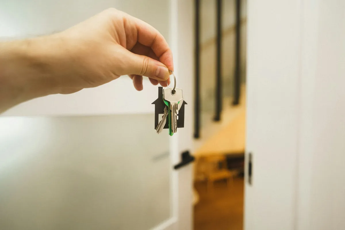 A hand holding a house-shaped keychain with new keys in front of a bright doorway, marking a first home milestone.